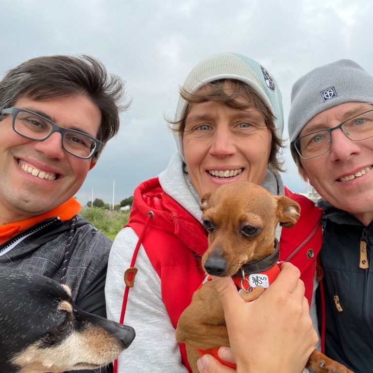 Das Team von WOVVE Drei Personen mit zwei Hunden lächeln in die Kamera, bei bewölktem Wetter.